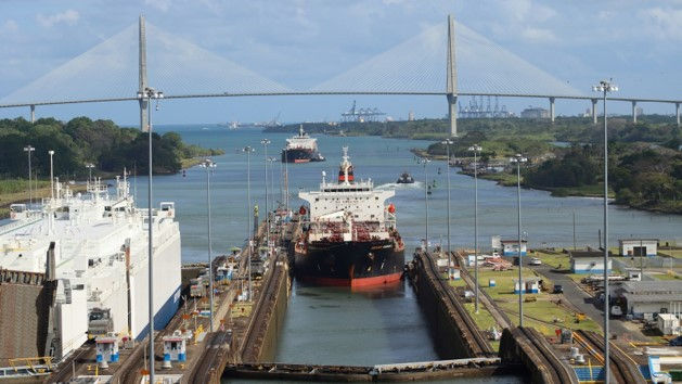 Cargo ships in a canal lock beneath a cable-stayed bridge, with port cranes and green islands under a cloudy sky.