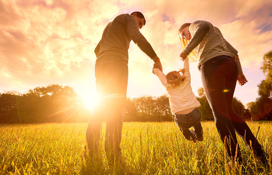 Parents swinging child in a sunlit field at sunset.