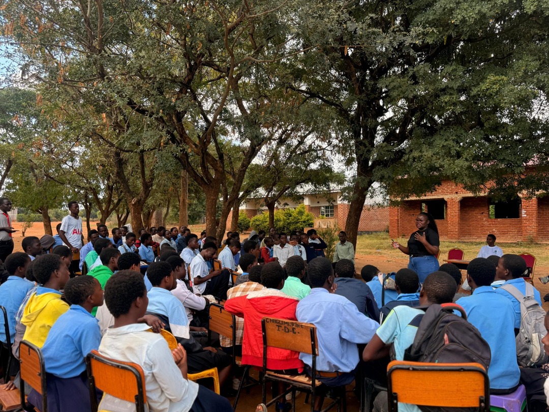Secondary students sit beneath trees at Chigumukile Community Day Secondary School while a Mthunzi Minds facilitator speaks.