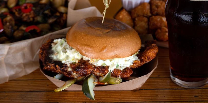 A fried chicken sandwich, with brussel sprouts and tater tots visible in the background.