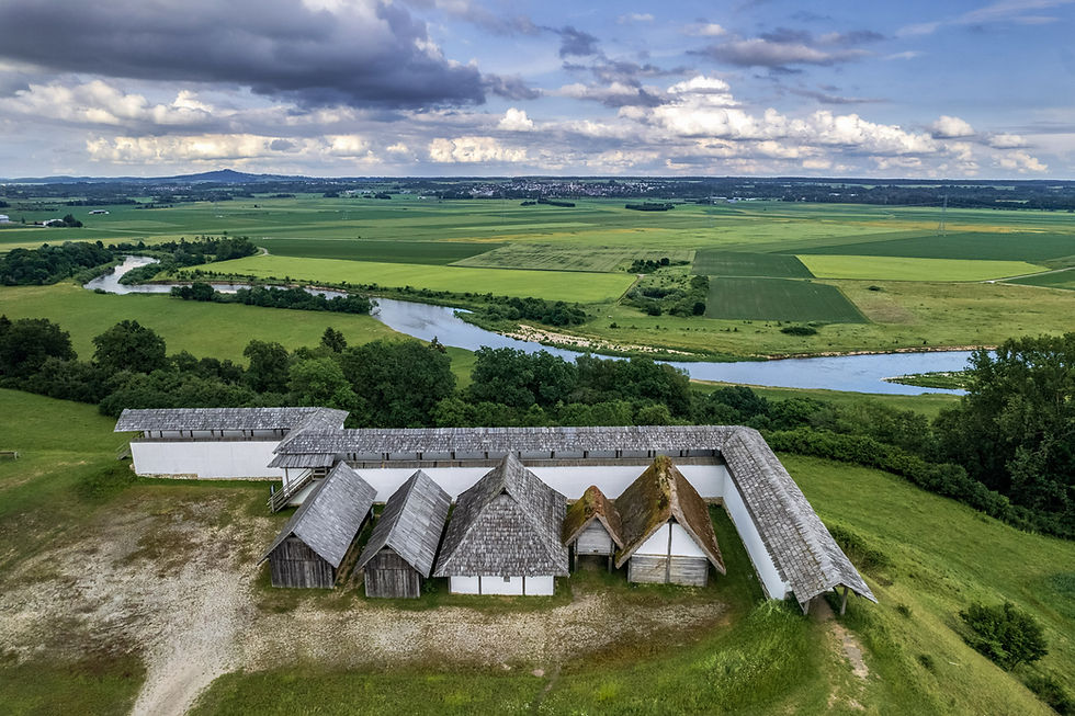 Luftaufnahme eines historischen Holzhauses mit Strohdach, umgeben von grünen Feldern und einem Fluss unter bewölktem Himmel. Friedliche Landschaft. Heuneburg