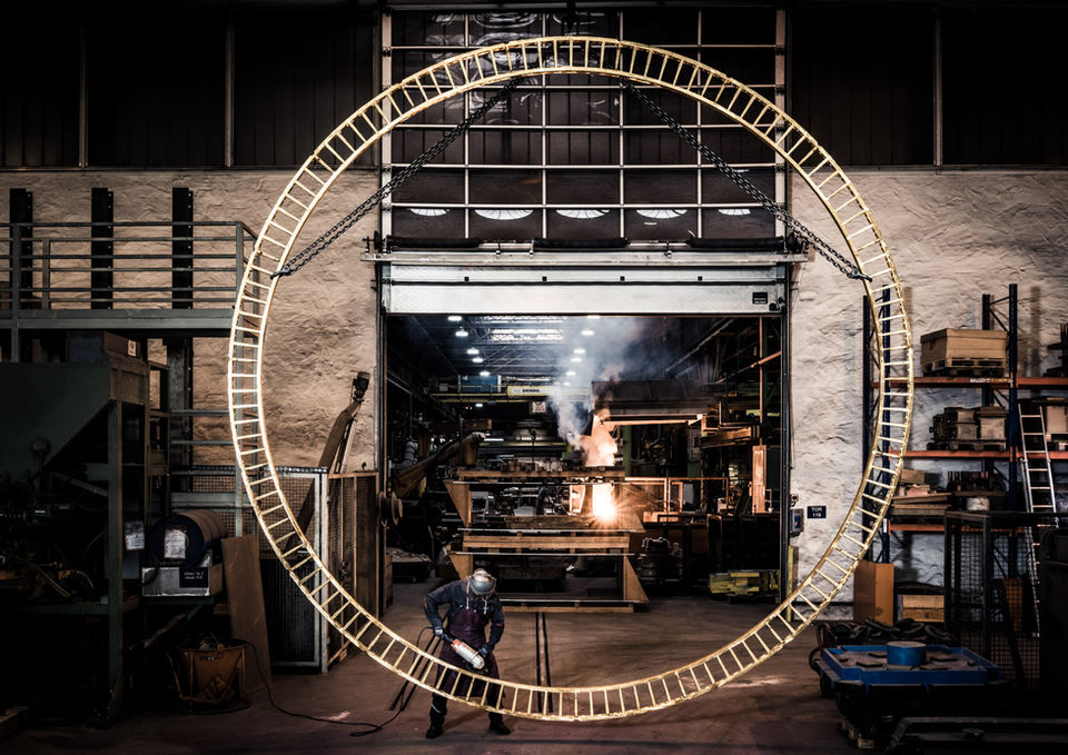 Gigantischer Metallring in dunkler Fabrikhalle mit Funkenflug und Arbeiter.