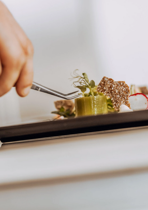 Koch garniert kunstvoll ein Gourmetgericht mit Pinzette auf schwarzem Teller.