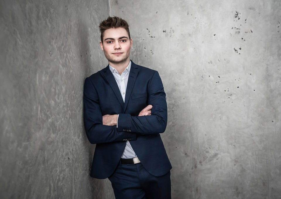 Junger Mann im Anzug lehnt an grauer Betonwand, Arme verschränkt.