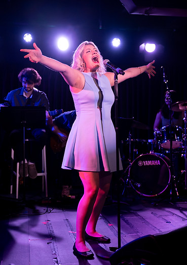 A blonde young woman in a blue dress singing enthusiastically on stage with outstretched arms