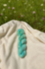 Skein of variegated soft greens and blue yarn with pops of yellow on a cream blanket in a field of grass and wildflowers