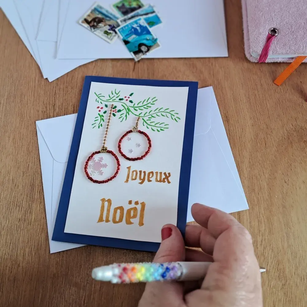 Carte joyeux noel bleue motif boules en perles posée sur un bureau avec une main qui l'ouvre