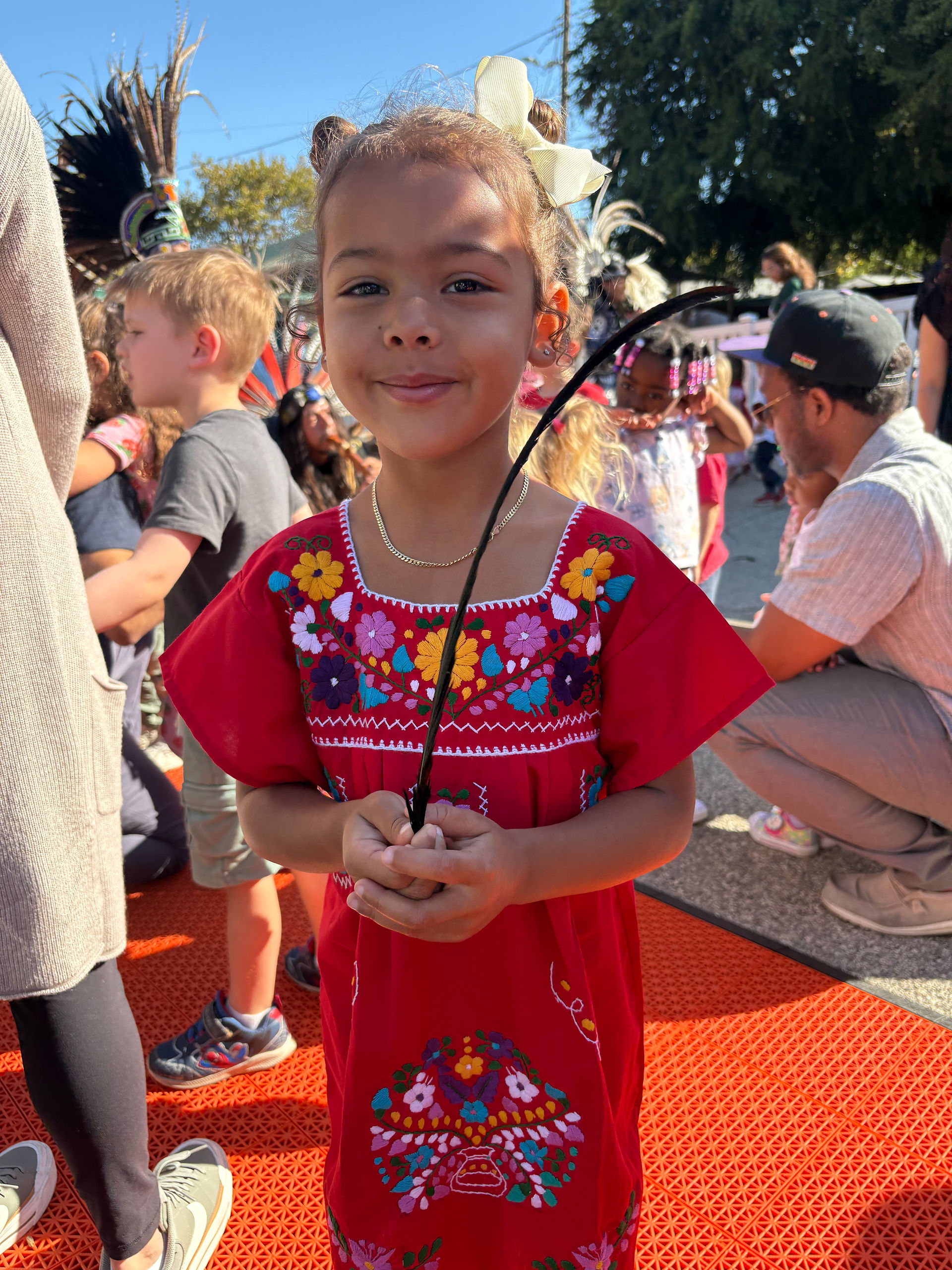 Young girl smiles in a red dress with embroidery, holding a feather.