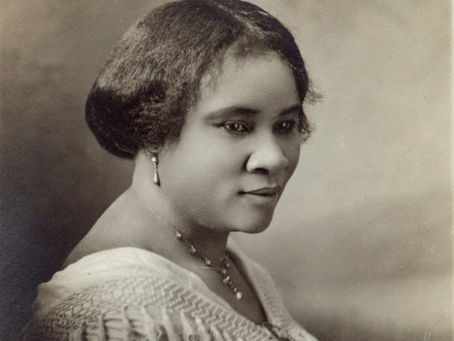 Portrait of a woman with styled hair and necklace, wearing a textured white blouse. She gazes softly to the side, set against a neutral background.