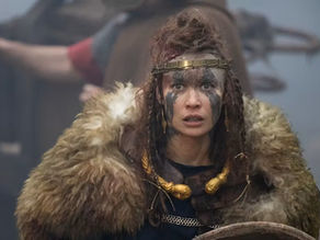 Warrior woman in fur cloak and face paint looks alert, holding a shield. Blurred background with weapon and foggy setting. Intense mood.