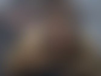 Warrior woman in fur cloak and face paint looks alert, holding a shield. Blurred background with weapon and foggy setting. Intense mood.