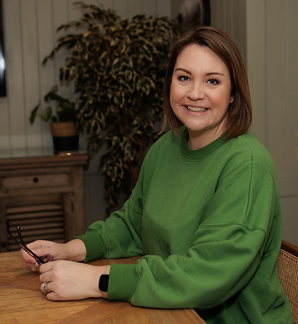 Woman in green jumper sat at a table holding glasses whilst looking at the camera smiling.