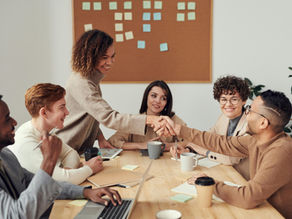 A diverse group of colleagues engaged in a positive and collaborative business meeting, sharing a handshake over a conference table filled with notebooks, coffee, and a laptop.