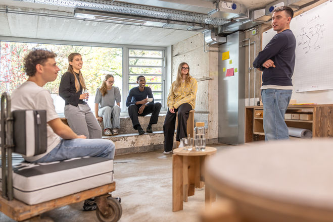 Ausschnitt aus einer Besprechungssituation, sechs Personen, männlich und weiblich, fünf Personen sitzen auf Besprechungsmöbeln, gebaut aus upgecycleten Materialien, eine Person steht vor einem Whiteboard, welches Bestandteil eines mobilen Besprechungswagen ist