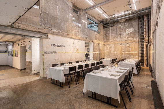 Untere Etage des Bilker Bunkers mit Tischen und Stühlen in einem Setup für Fine Dining, insgesamt vier Tafeln, die längs im Raum stehen, eingedeckt mit weißen Tischdecken, Tellern, Servietten und Weingläsern