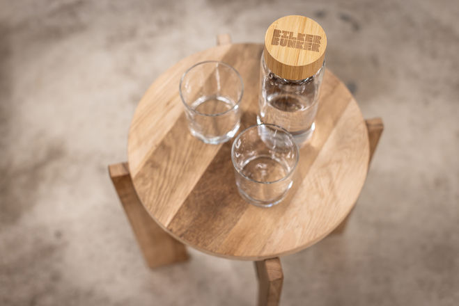 Zu sehen ist eine Wasserflasche für Mineralwasser, welche mit zwei Gläsern auf einem selbstkonstruiertem Stuhl steht. Dieser wurde aus recycletem Material gebaut.