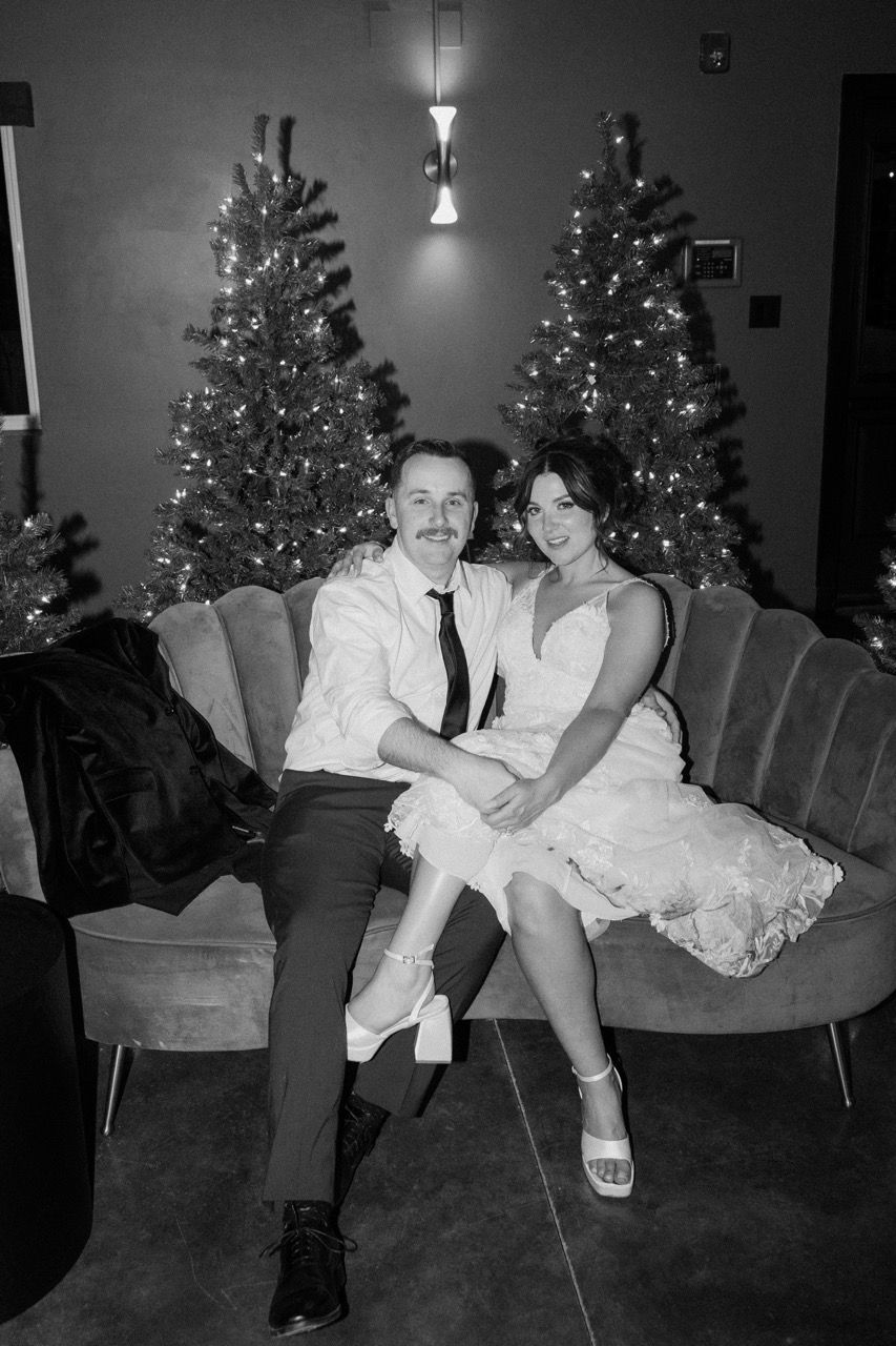 a bride and groom sit on a couch in front of christmas trees