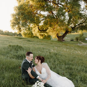 a bride and groom sit in a grassy field under a tree