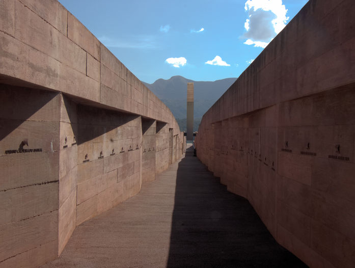 A imagem mostra um corredor entre dois muros de concreto com vários nomes gravados, cada um representando uma vítima. Ao fundo, há um monumento alto e no horizonte vê-se montanhas e céu azul.