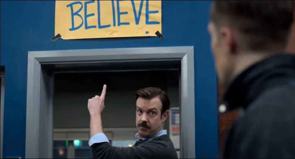 Ted Lasso pointing to yellow sign that says Believe
