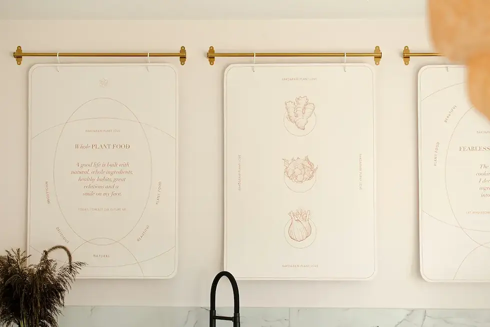 Oversized botanical prints hung vertically on brass rod above kitchen sink as modern gallery wall alternative to frames
