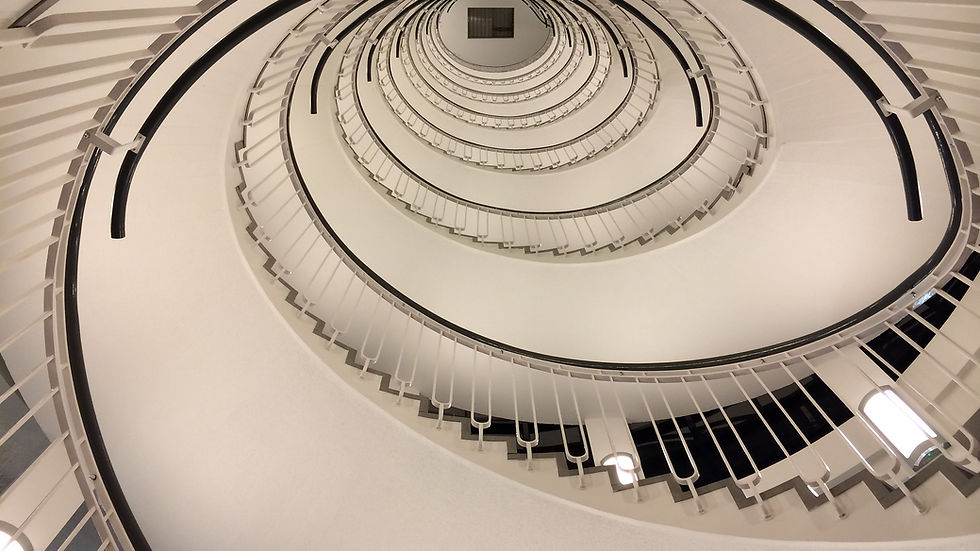 Looking up at a spiral staircase with white railings and beige walls, creating a hypnotic pattern. Bright lights illuminate the scene. Representing spiral of anxiety worry and avoidance