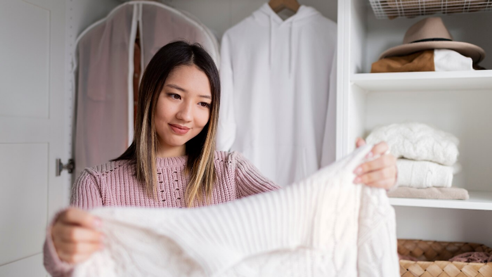 Woman in a tidy closet holds up a white knit sweater, smiling softly, with white shirts, hats, and baskets behind her.