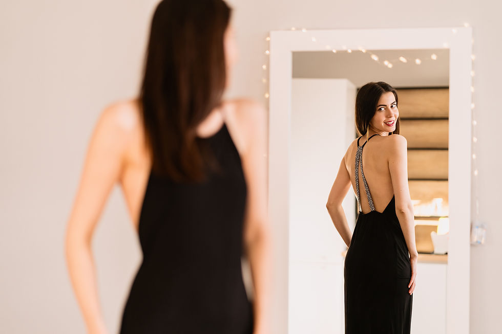Woman in a black backless dress smiles at her reflection in a mirror strung with warm fairy lights.