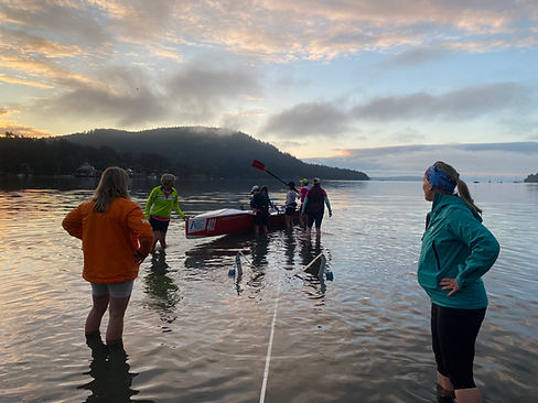 Red coastal quad being walked by several athletes into shallow pre-dawn water with a misty tree-covered hill in the background. Clouds lit up with warm colours of sunrise.