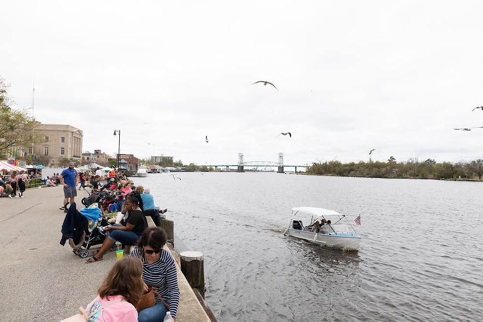 People sit along a the Riverwalk in Wilmington under a cloudy sky. A small boat with an American flag navigates the river, with a bridge in view.