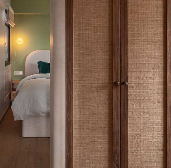 HDB master bedroom corridor with walnut and woven grasscloth island wardrobe in foreground and sage green bedroom wall with upholstered bed beyond
