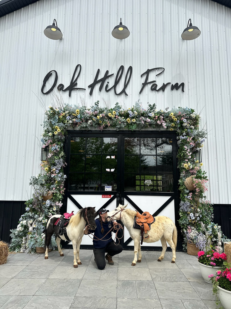 Person kneeling between two ponies in front of the Oak Hill Farm barn with floral-decorated doorway GEO targeted | Oak Hill Farm, Holmdel, NJ