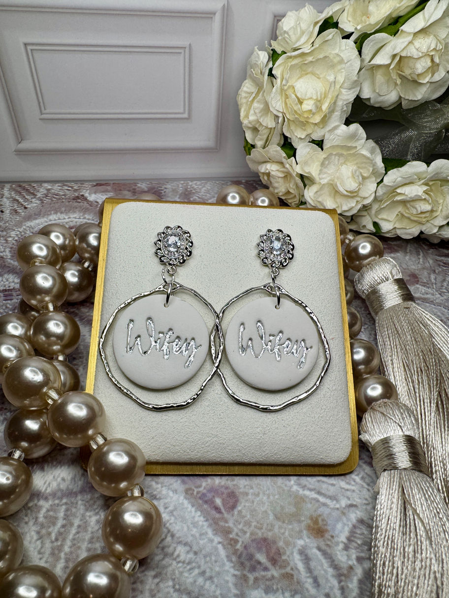 White “Wifey” earrings with silver foil lettering on a neutral display, with pearls and white florals in the background.