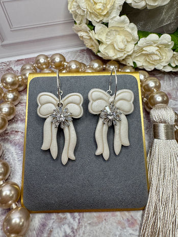 Cream bow earrings with white gold hoops and crystal center accents on a gray velvet display.