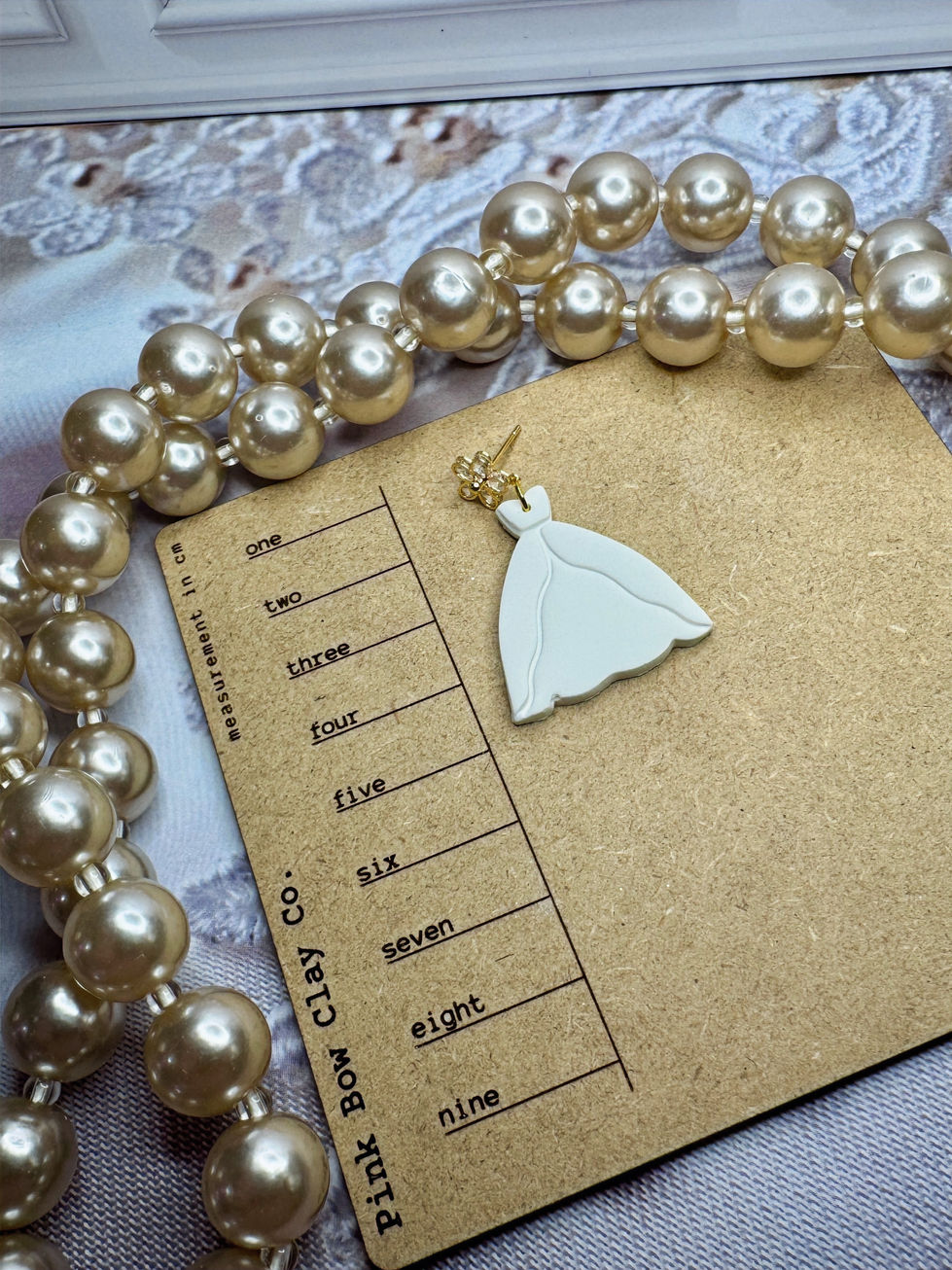 Single white bridal gown–shaped clay earring on a measurement card with gold floral CZ stud.