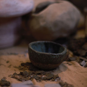 Small glazed pottery bowl sits on sand covered surface surrounded by broken pottery shards and larger vessels in the distance.