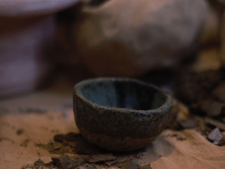 Small glazed pottery bowl sits on sand covered surface surrounded by broken pottery shards and larger vessels in the distance.