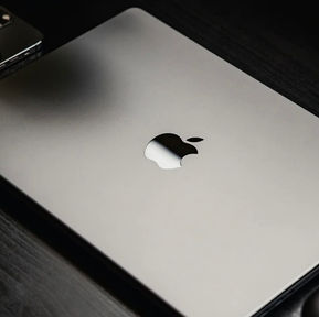 Ordinateur portable Apple MacBook en aluminium fermé, posé sur un bureau sombre avec un iPhone partiellement visible à côté.