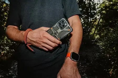 Homme en T-shirt noir tenant un smartphone à coque robuste, bracelet orange et montre, dans une forêt sombre.