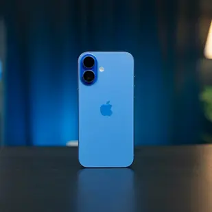 iPhone 16 bleu posé verticalement sur une table, avec un éclairage d’ambiance flou en arrière-plan dans un intérieur moderne