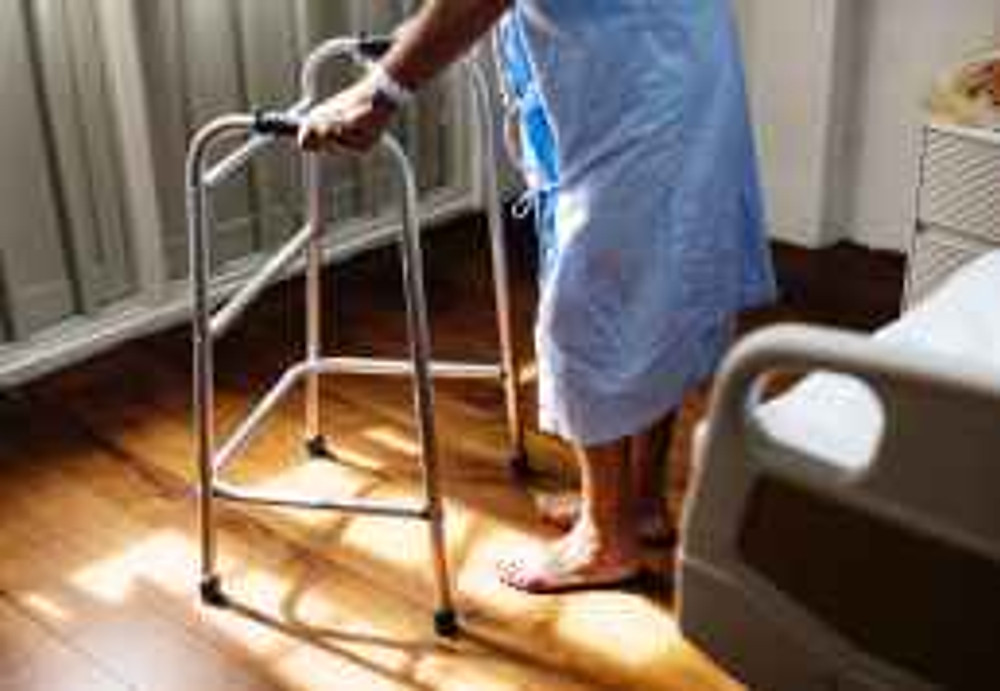 person in hospital gown using walking frame beside hospital bed