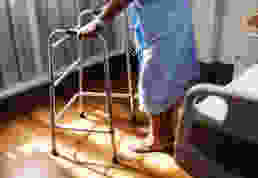 person in hospital gown using walking frame beside hospital bed