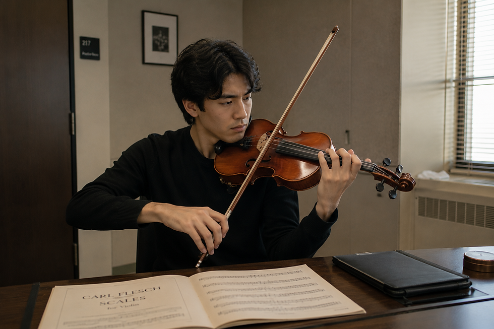 Man playing violin intently, seated at a table with sheet music labeled "Carl Flesch Scales." Room has neutral tones and soft lighting.