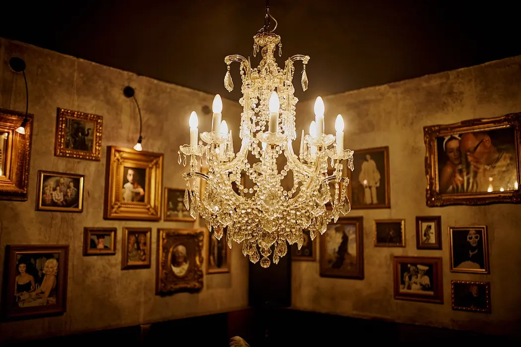 A glittering crystal chandelier in front of an array of gilt-framed pictures on a wall.