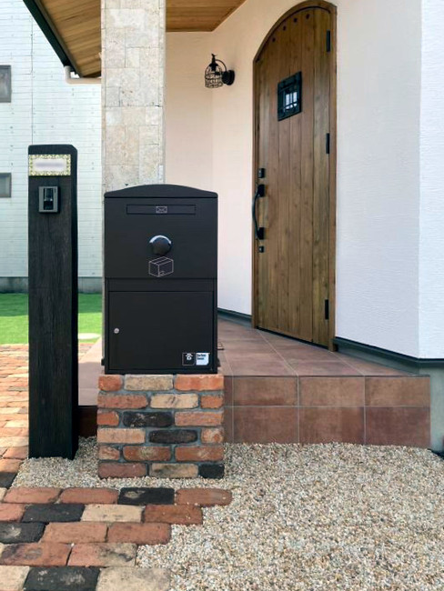 戸建て玄関前に設置されたブラウンの大容量宅配ボックス。レンガ調の台座に設置され、デザイン性と実用性を兼ね備えています。