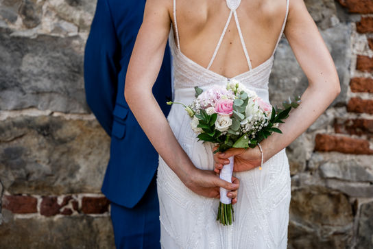 Detailfoto Brautrücken und Brautstrauß, die vor ihrem Bräutigam und einer Backsteinwand steht.
