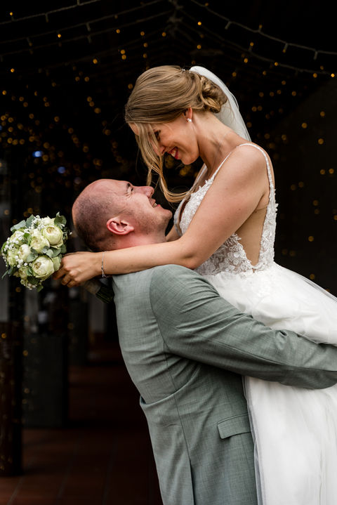 Bräutigam hebt Braut hoch, Braut hält Brautstrauß hinter seinem Kopf fest.