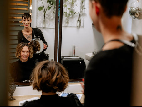 Das Bild zeigt eine Friseurin und eine Kundin in einem Salon. Zu sehen ist die Kundin im Spiegel und die Friseurin dahinter. Die Friseurin zeigt der Kundin mit einem Spiegel von hinten ihre Frisur.