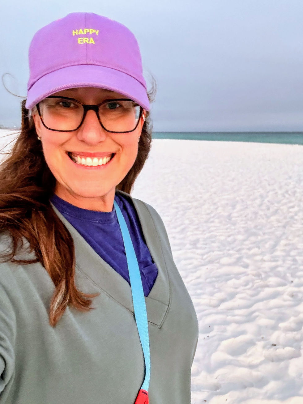 Woman smiling on the Emerald Coast beach wearing a purple Happy Era hat, overcast sky and white sand behind her.