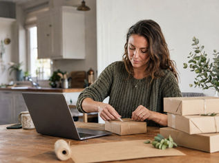 A woman packing parcels at her kitchen table beside a laptop, running a small online side hustle from home.